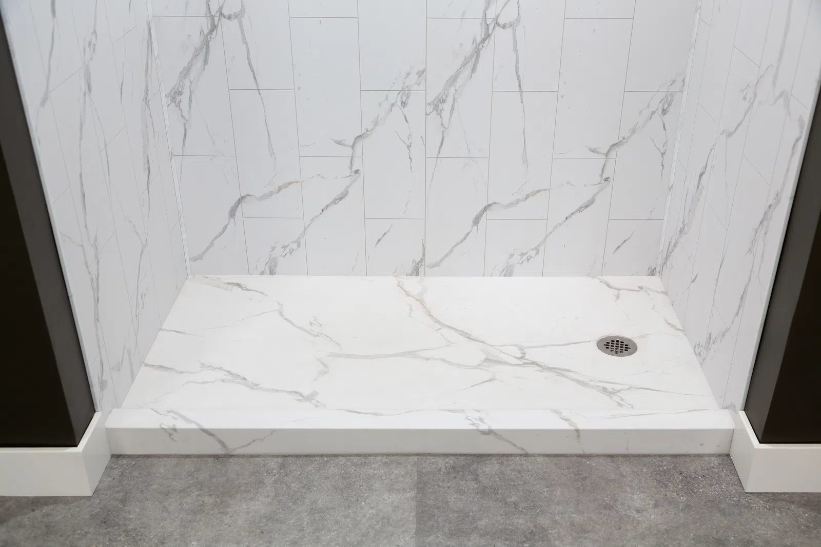 Modern shower area with white marble tile walls and matching floor, featuring a metal drain and gray surrounding floor.