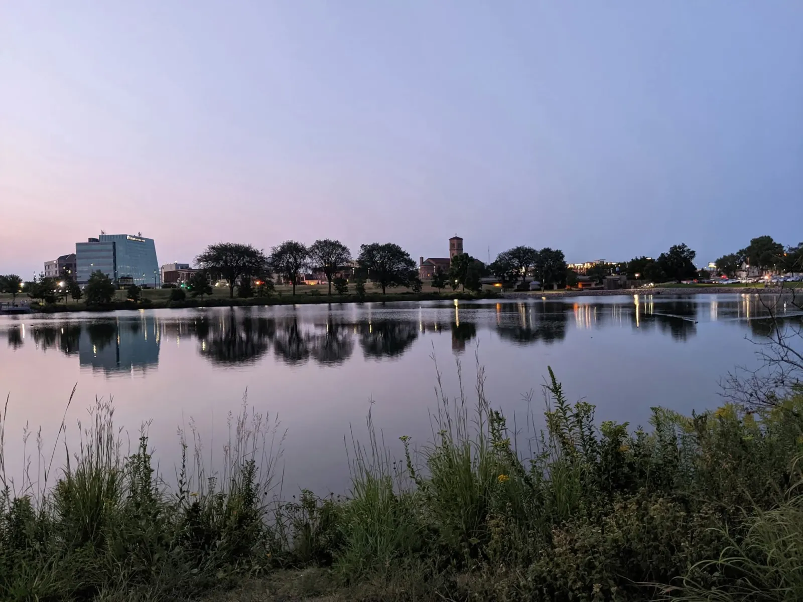 Calm lake reflecting trees and city buildings under a clear twilight sky with foreground greenery.