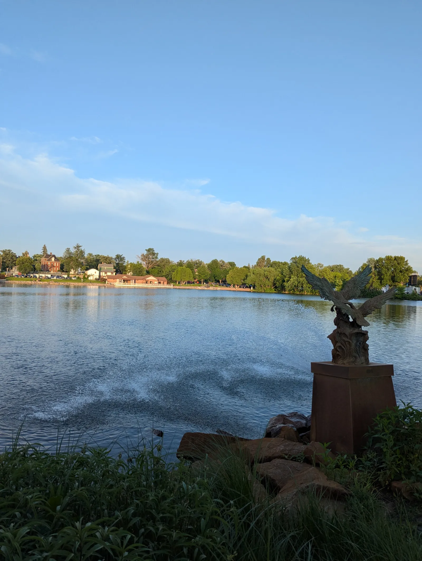 Eagle statue overlooking a calm lake with trees and houses under a clear blue sky at sunset.