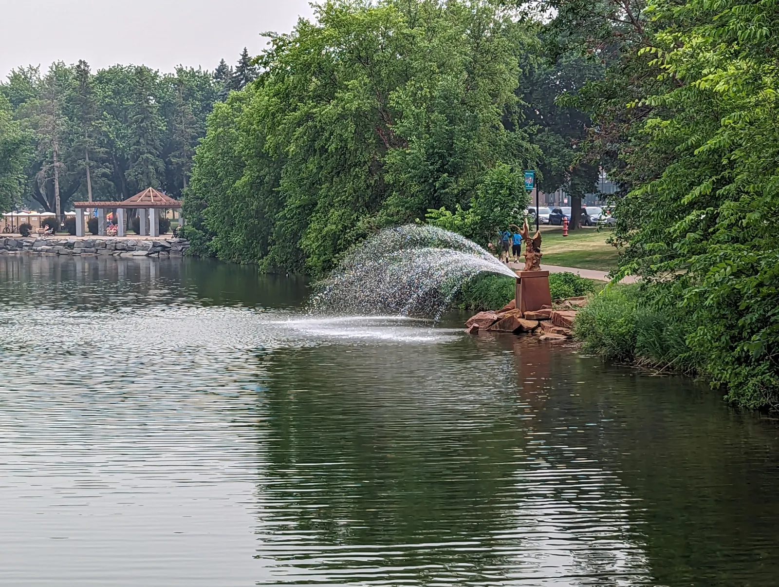 Fountain spraying water into a calm lake surrounded by lush green trees and a walking path beside it.
