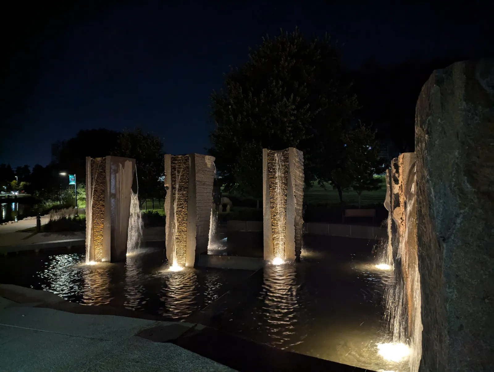 Illuminated stone pillars with cascading water in a nighttime outdoor fountain surrounded by trees and pathways.
