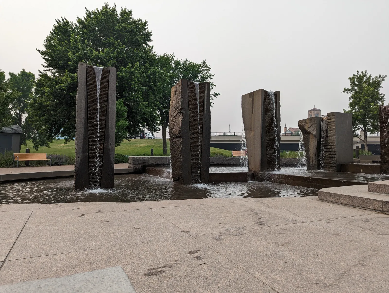 Modern stone pillars with waterfalls in a park fountain surrounded by trees and benches on a cloudy day