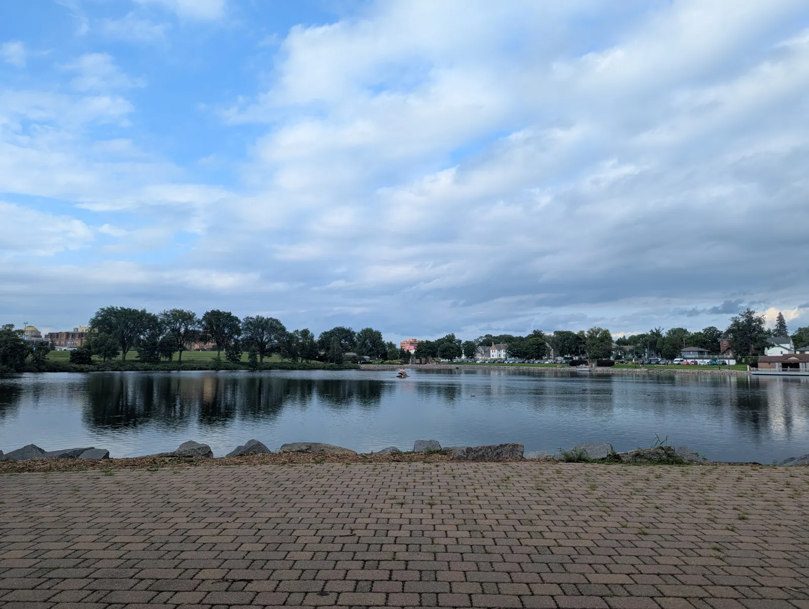 Calm lake with reflections of trees and cloudy sky viewed from a brick-paved shoreline in a park.