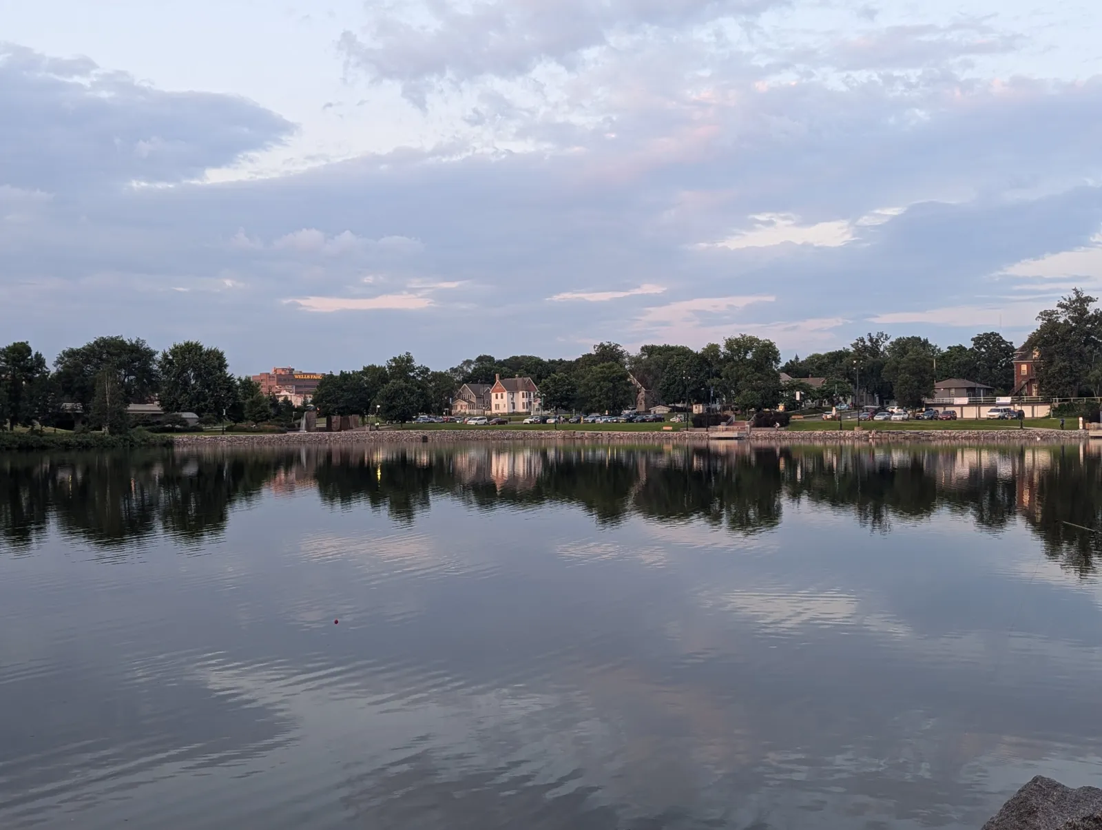 Calm lake reflecting trees and houses under a cloudy sky during evening light.