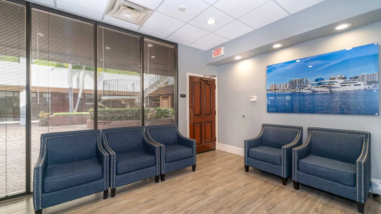 Modern waiting room with blue upholstered chairs, wood flooring, large window blinds, and a yacht photo on the wall.