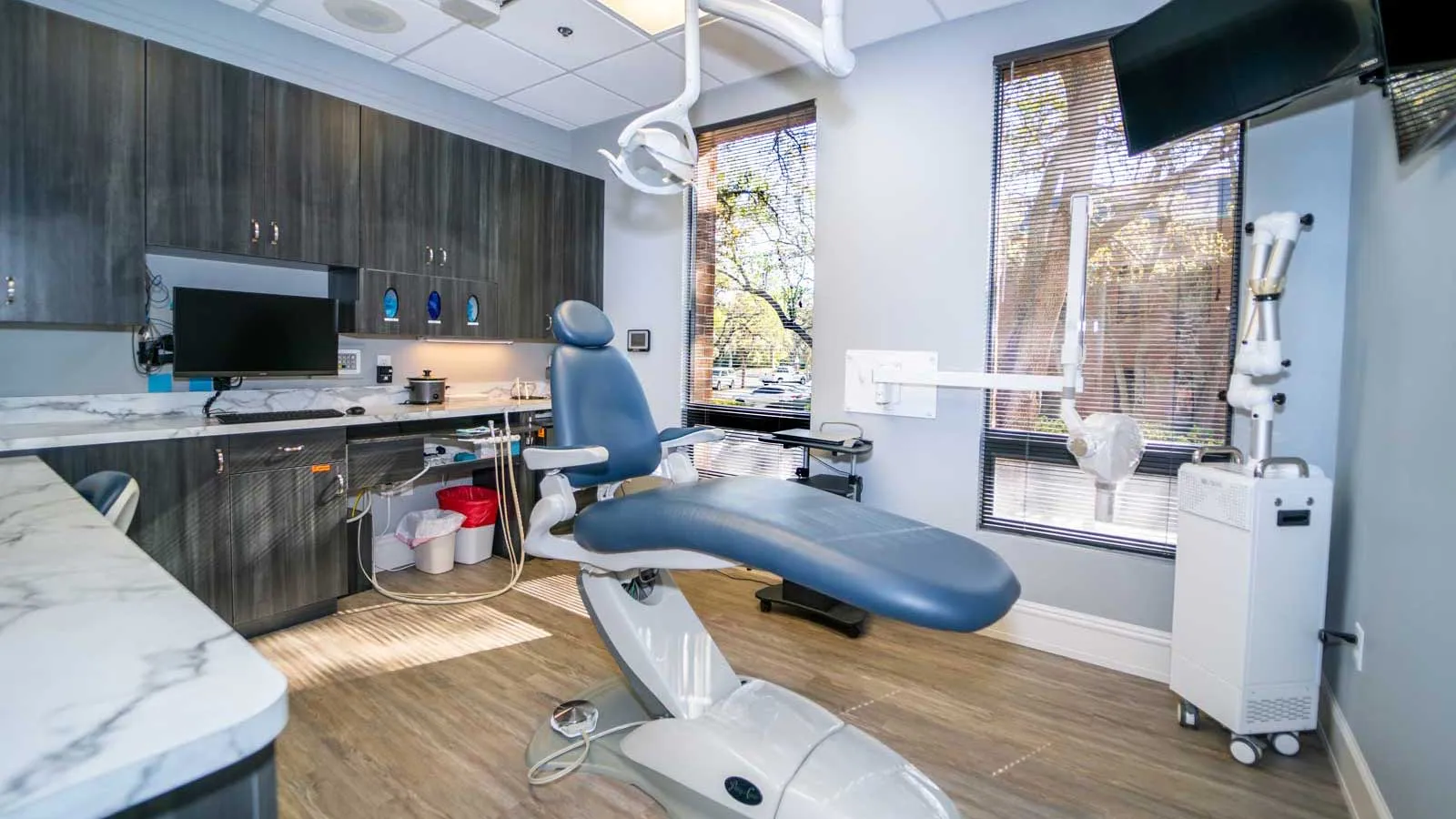 Modern dental office with blue patient chair, computer, large windows, and wooden cabinets under bright lighting.