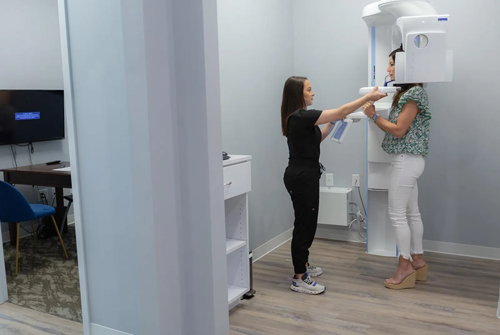 Dental technician assists patient in a floral shirt and white pants during a panoramic X-ray scan in a clinic.