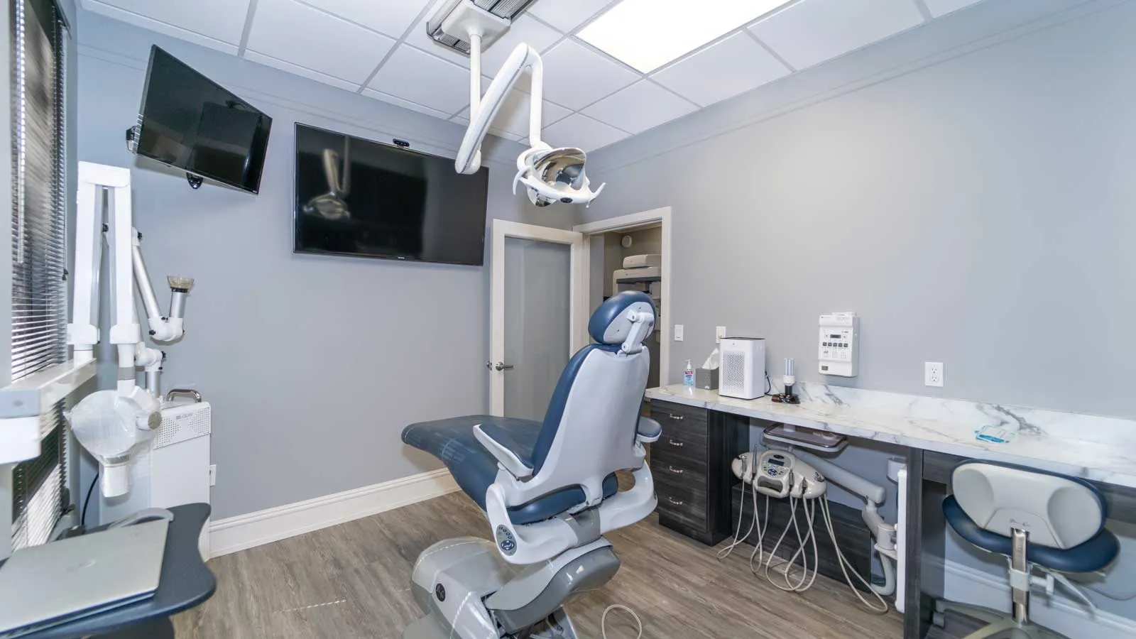 Modern dental office with patient chair, overhead light, monitors, and dental equipment on wood flooring.
