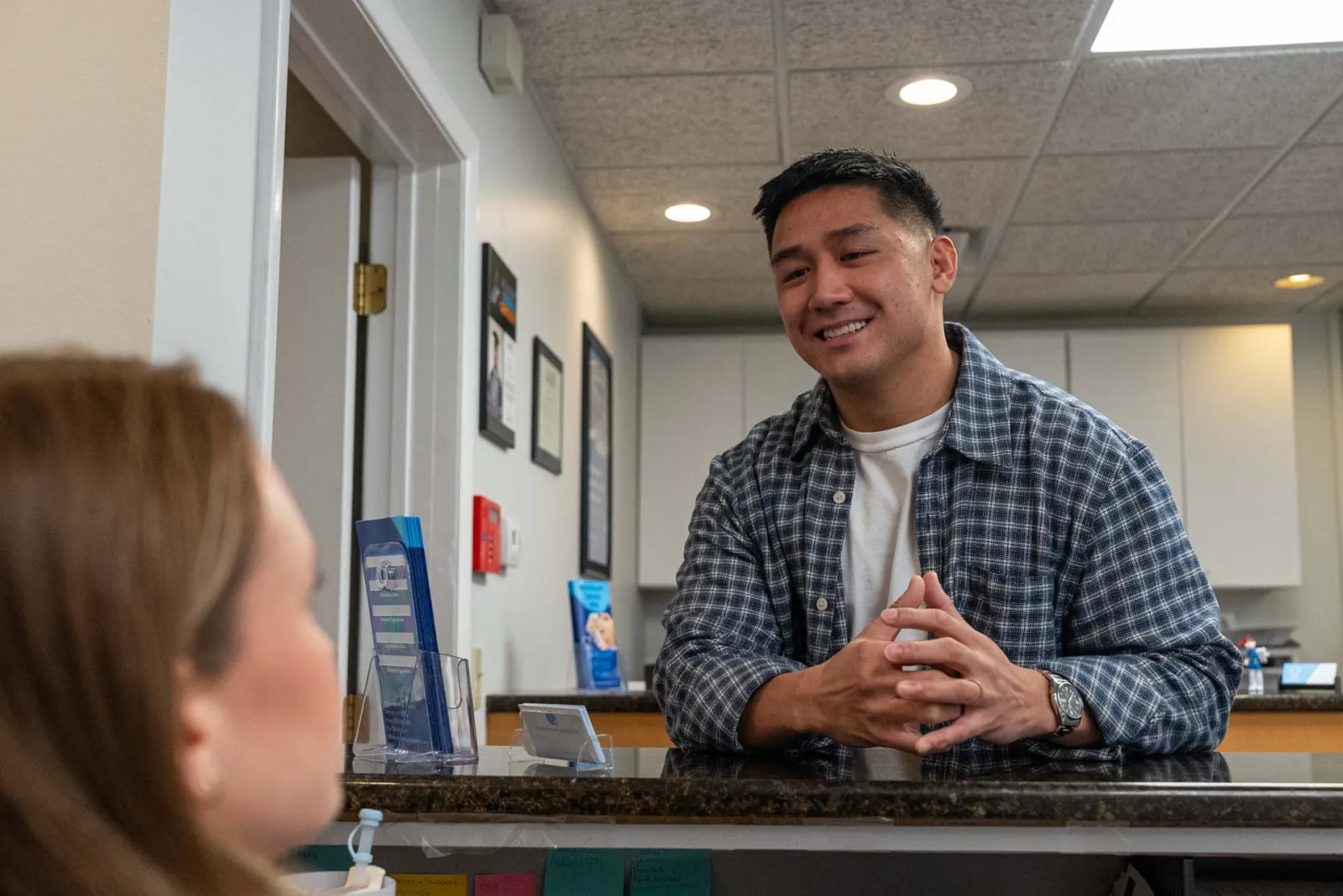 Smiling man in plaid shirt talks to a woman across a reception desk in a professional office setting.