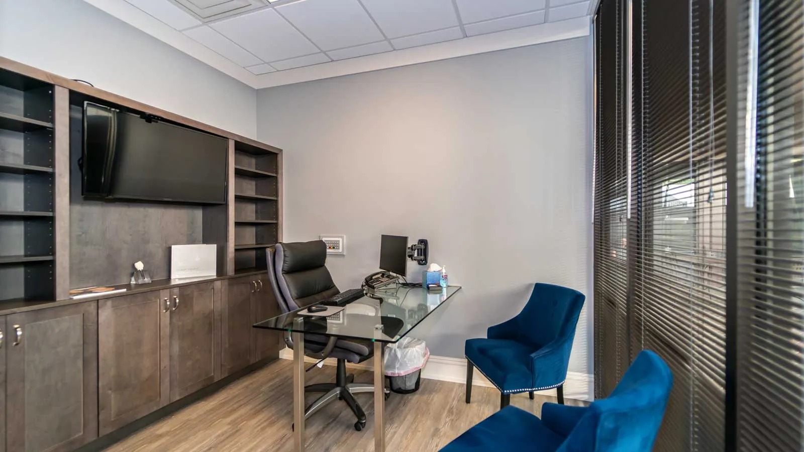 Modern office with glass desk, black swivel chair, blue guest chairs, and large TV on wood shelves.