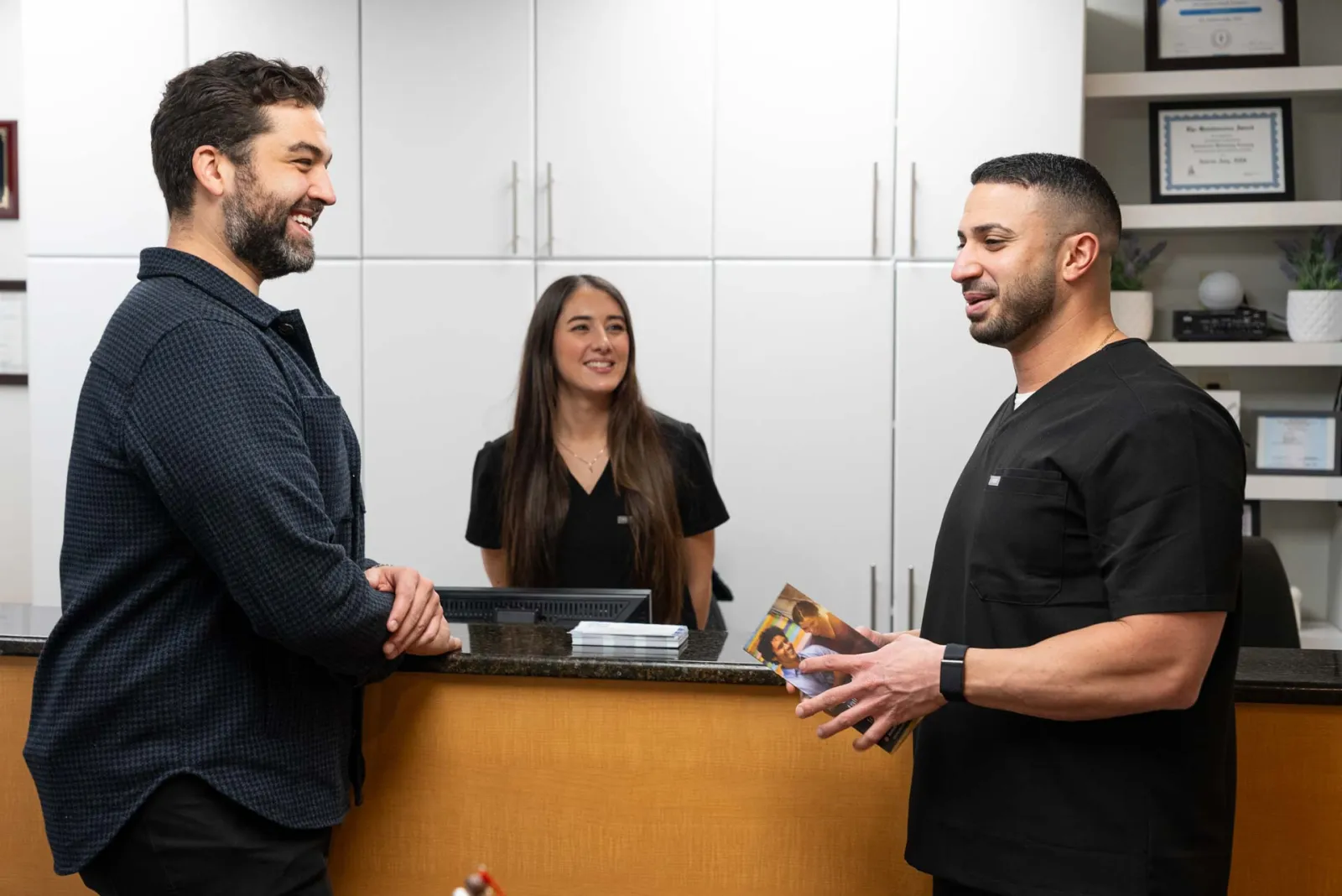 Two men speaking at a dental clinic reception desk with a female staff member smiling behind the counter