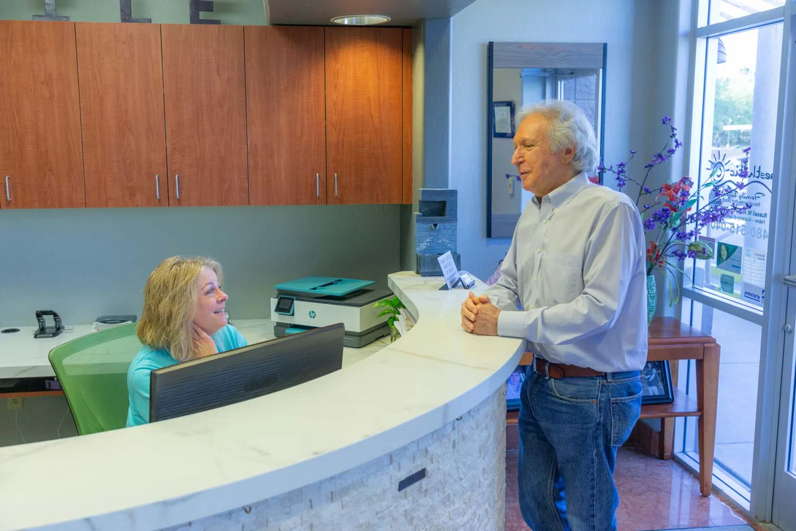 Senior man talking to receptionist at medical office front desk in bright waiting area