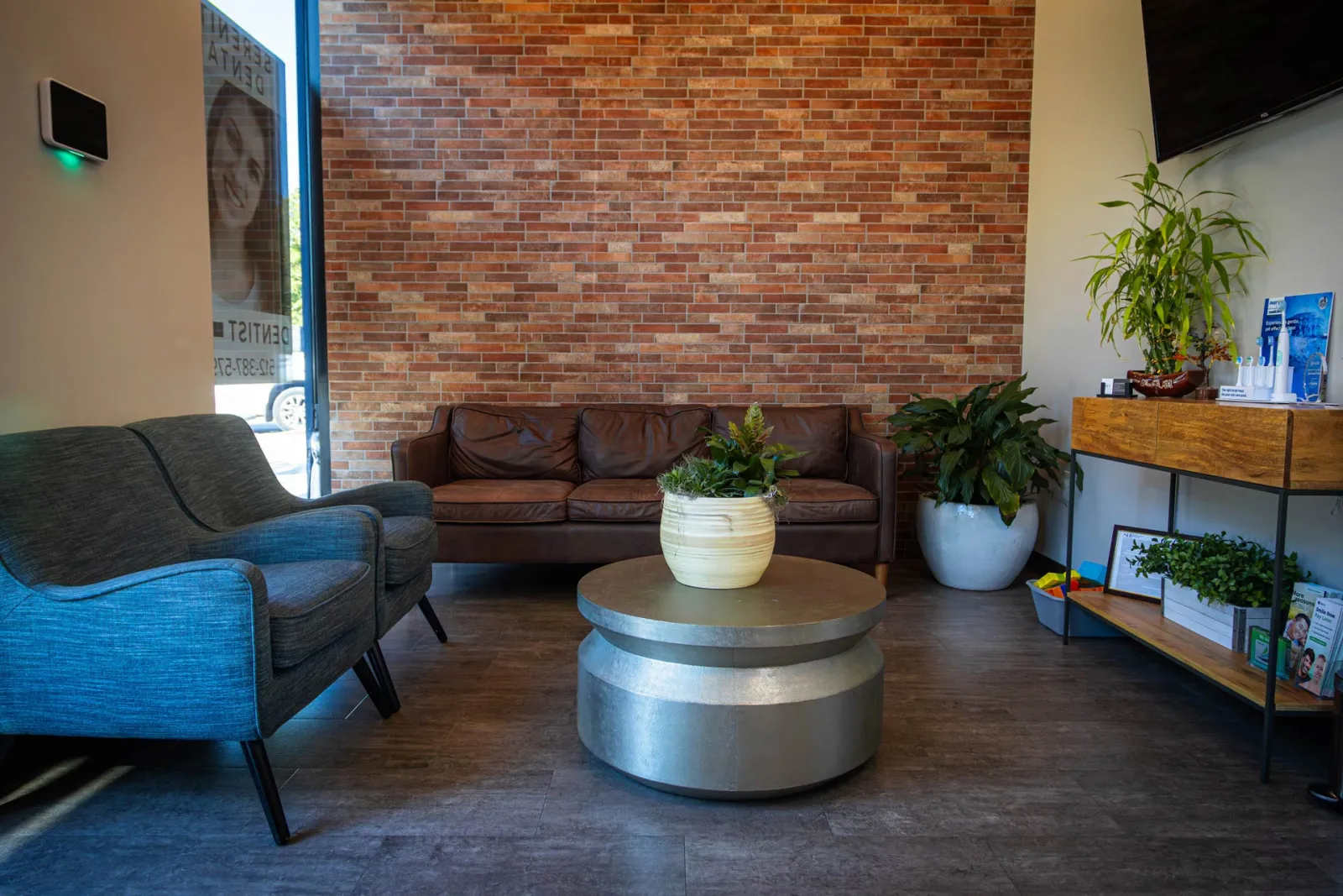 Modern waiting room with brown leather sofa, blue armchair, metal coffee table, and plants against brick wall.