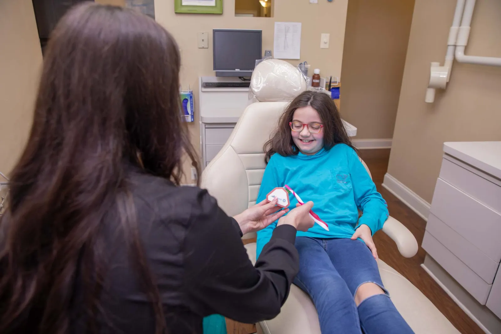 Dentist demonstrating proper tooth brushing technique to smiling girl sitting in dental chair at clinic.