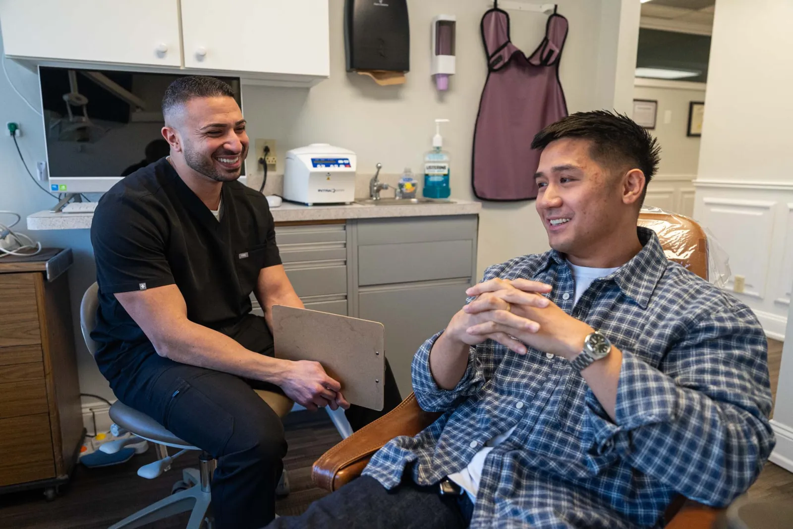 Dentist in black scrubs consulting with a smiling male patient sitting in a dental chair in a clinic.