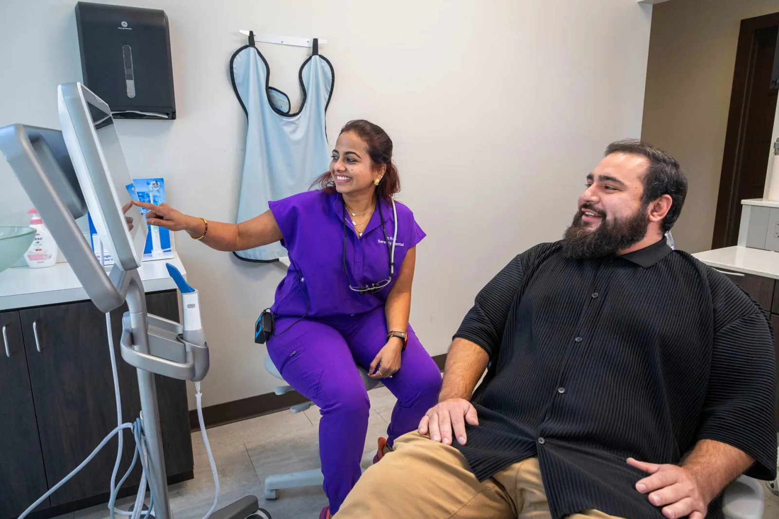 Female medical professional in purple scrubs showing a patient information on a monitor in a clinic room.