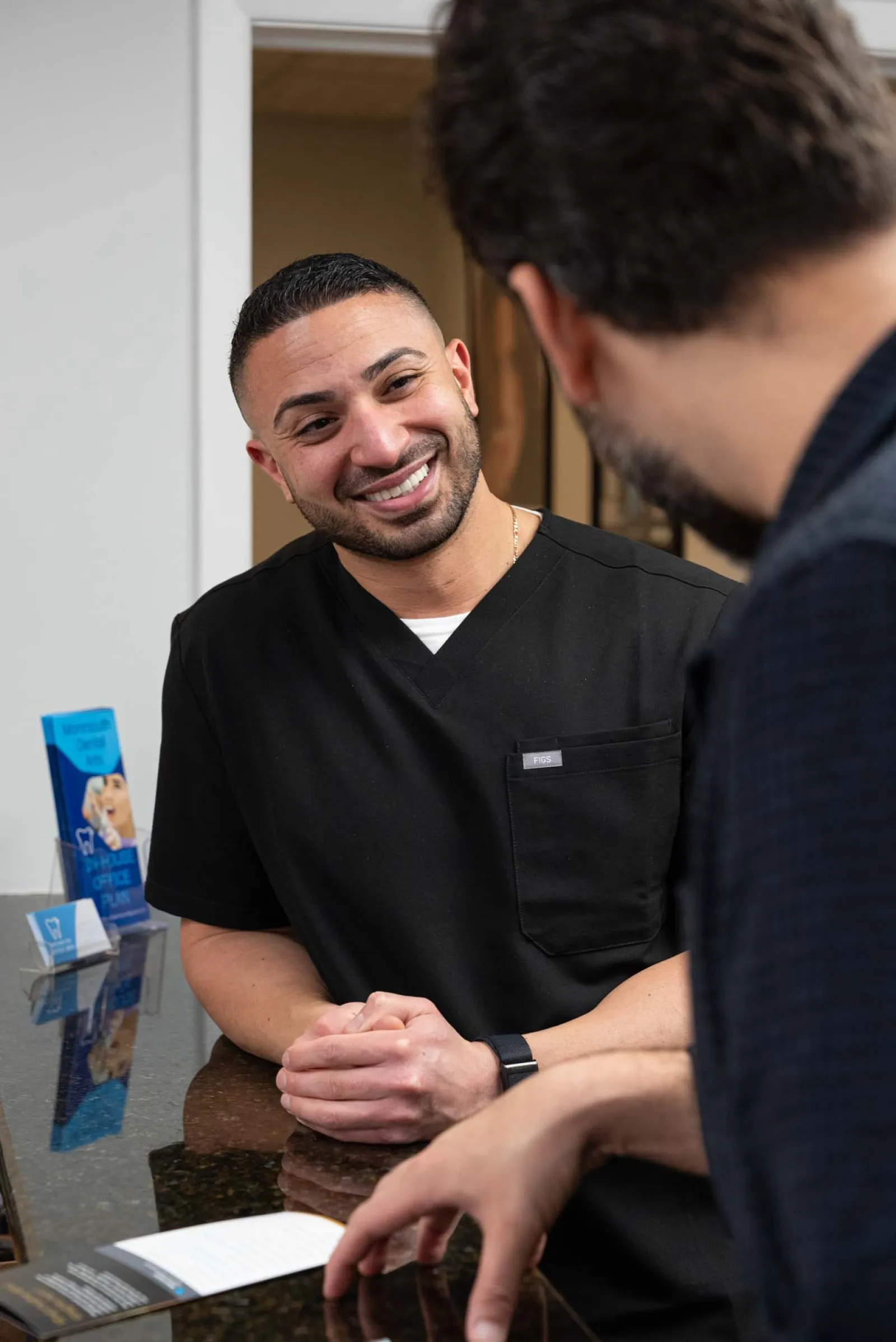 Smiling male healthcare professional in black scrubs talking with a patient across a desk in a medical office.
