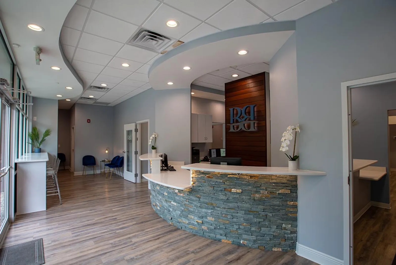 Modern dental office reception area with curved stone desk, wood flooring, blue chairs, and soft lighting.