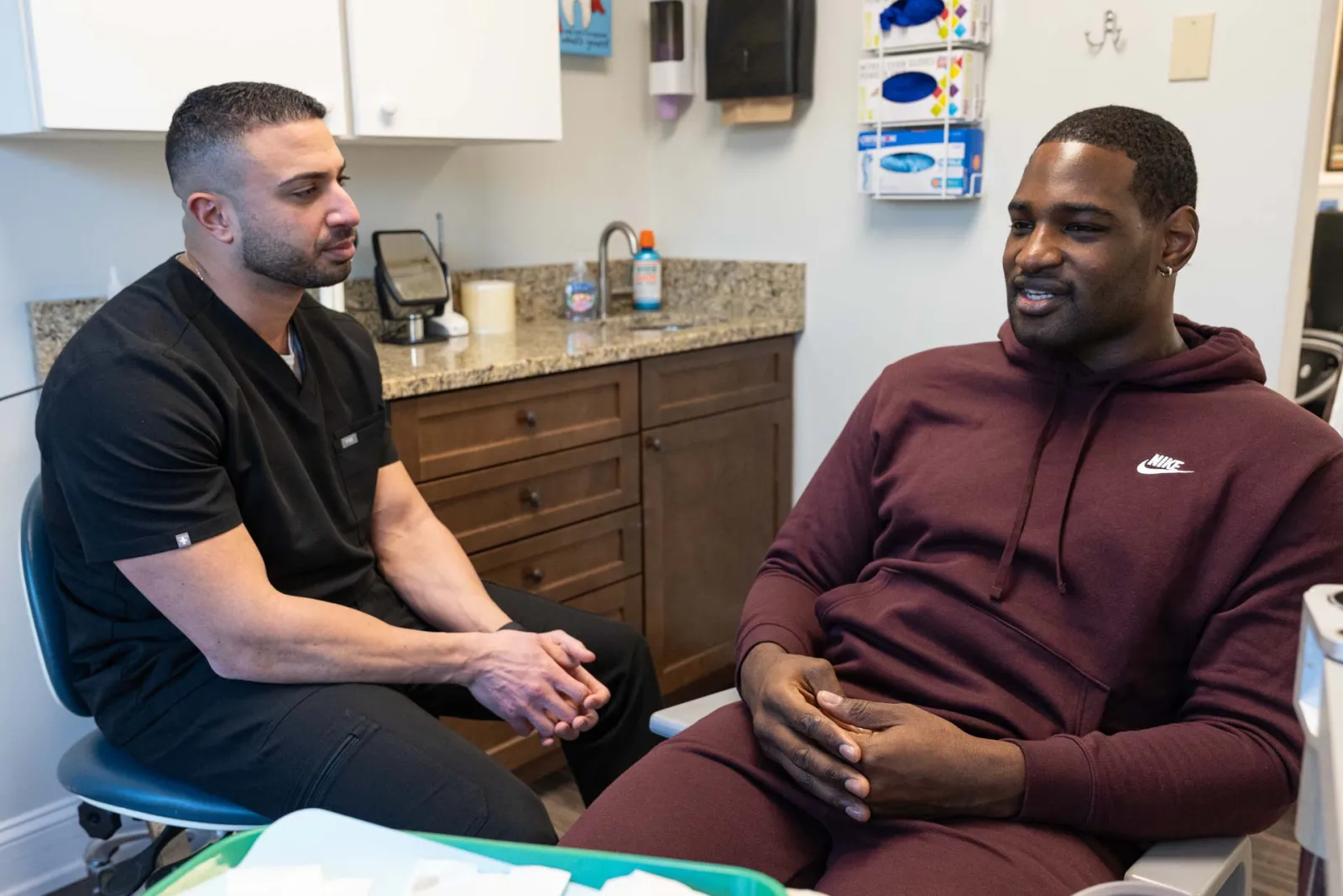 Dentist in black scrubs consulting a male patient in a burgundy Nike hoodie in a modern dental office.