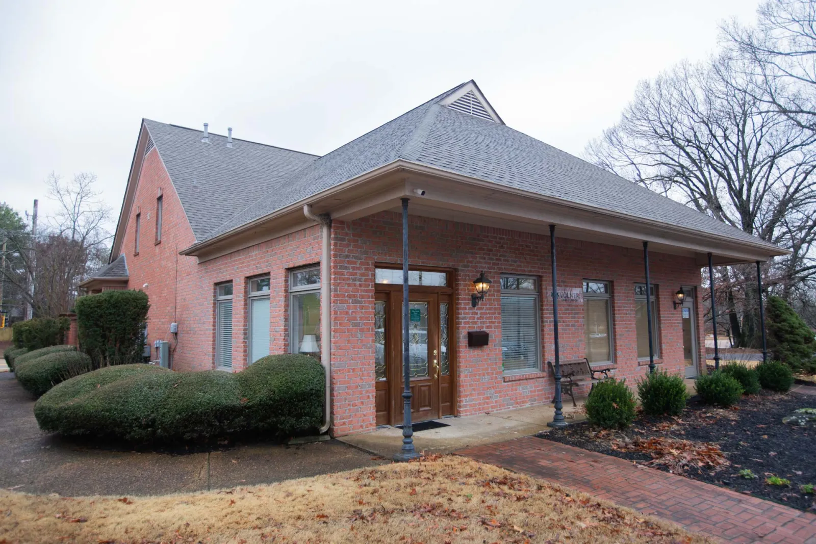 Single-story red brick building with peaked roof and large wooden door surrounded by bushes and a paved walkway.
