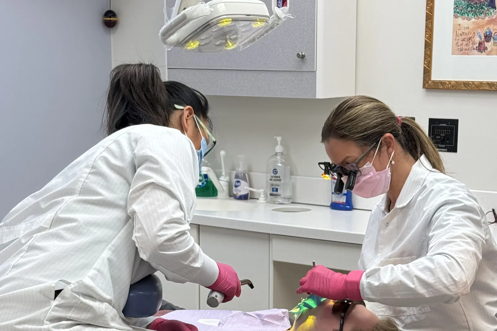 Two dentists wearing masks and gloves perform a dental procedure on a patient in a clinic room.
