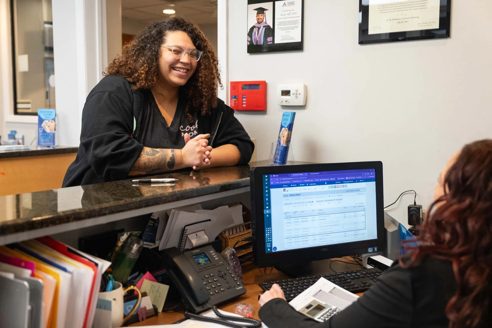 Smiling woman with curly hair talks to receptionist at office counter with computer and phone.