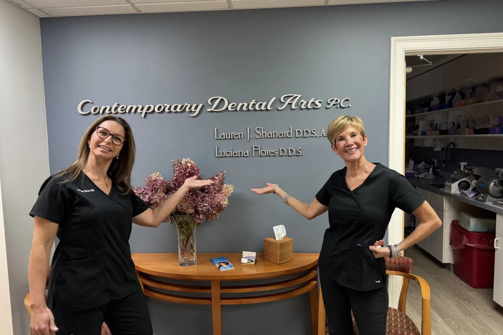 Two smiling female dentists in black scrubs pose in front of Contemporary Dental Arts P.C. sign with flowers and brochures.