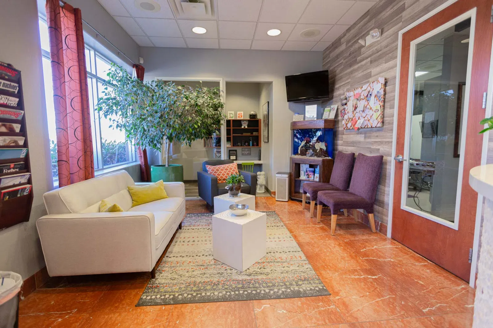 Bright modern waiting room with beige sofa, purple chairs, large plant, TV, and decorative fish tank on wooden floor.