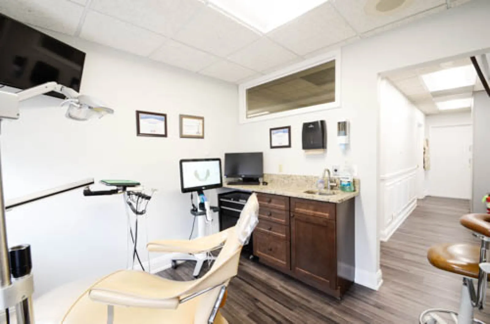 Bright dental office with beige patient chair, monitors, cabinets, and certificates on the wall.