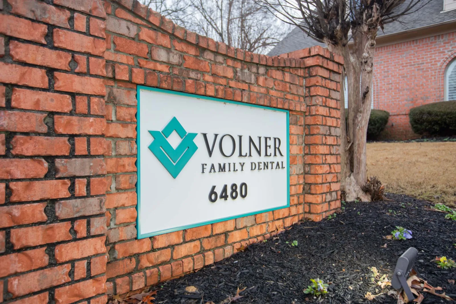 Brick sign for Volner Family Dental with teal logo and address 6480 in a landscaped yard.