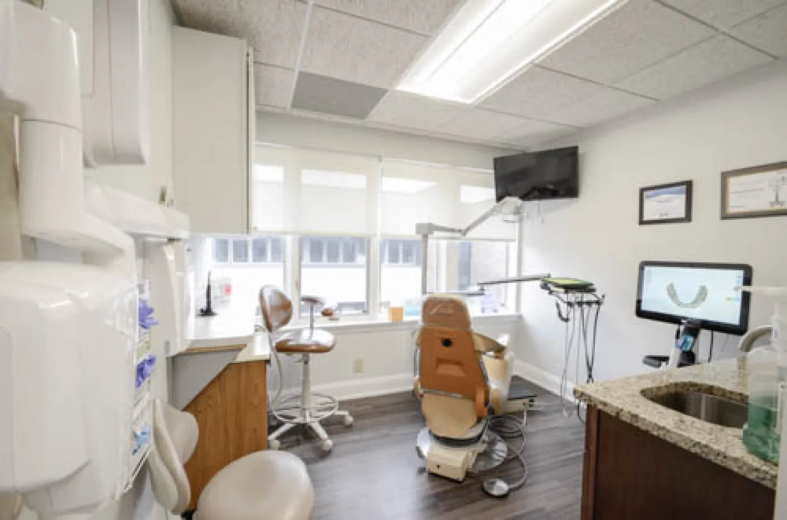 Bright dental office with patient chair, monitor displaying dental scan, stools, and large windows providing natural light