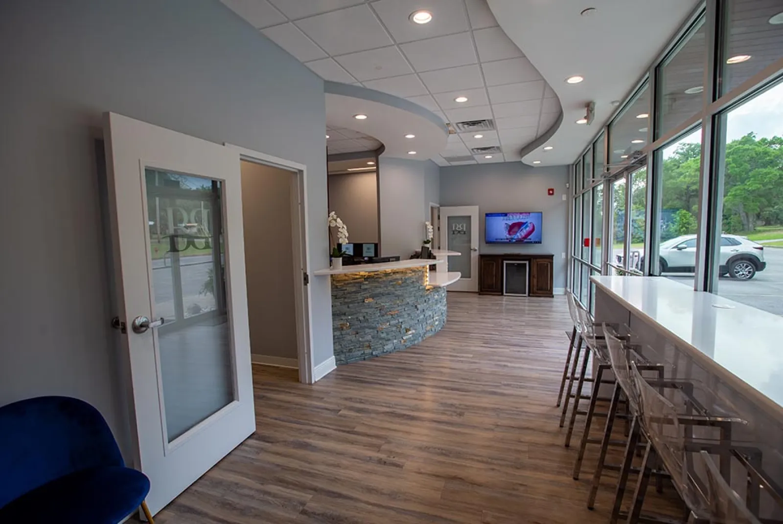 Modern dental office reception area with curved stone desk, wood floor, and large windows with bar stools.