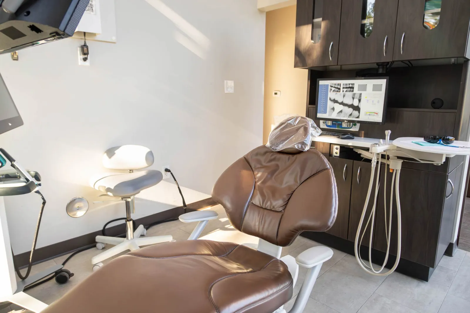 Modern dental clinic with a brown leather dental chair, monitor displaying dental X-rays, and equipment.