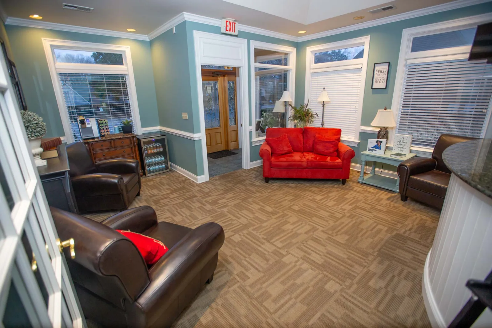 Cozy waiting room with red sofa, brown leather chairs, potted plants, lamps, and large windows with blinds.
