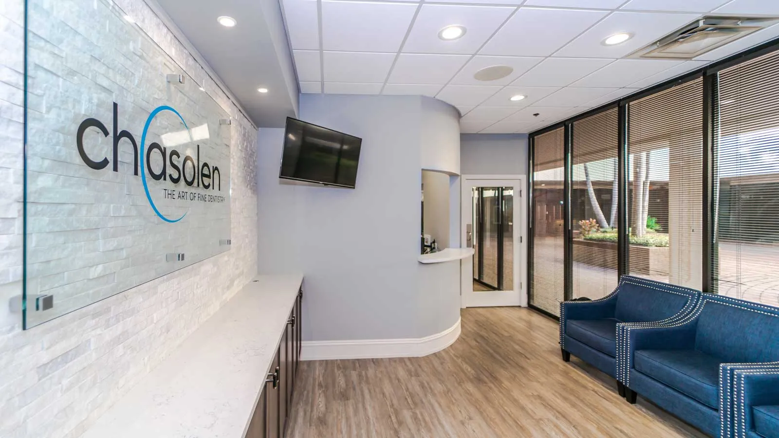 Modern office lobby with blue chairs, large windows, wall-mounted TV, and Chasalen logo on white brick wall.