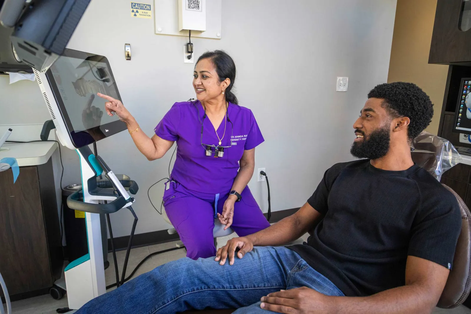 Female dentist in purple scrubs explaining dental scan results to male patient in clinic chair