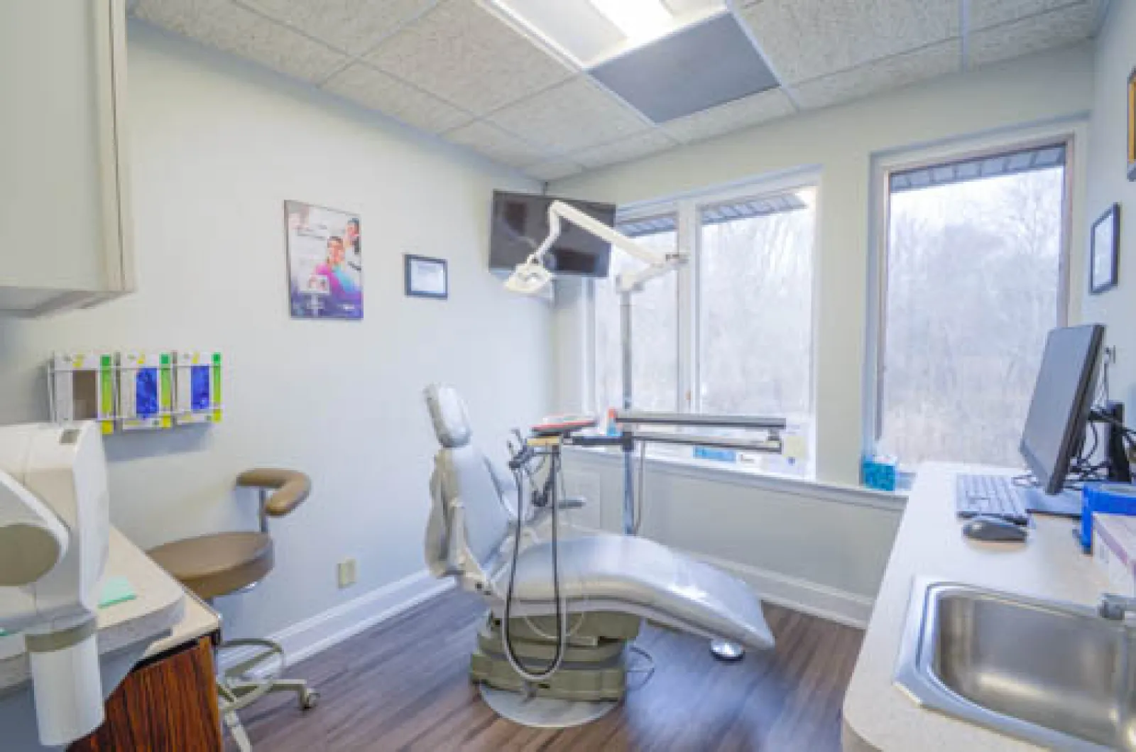 Bright dental clinic room with modern chair, equipment, large window, and clean wooden floor.