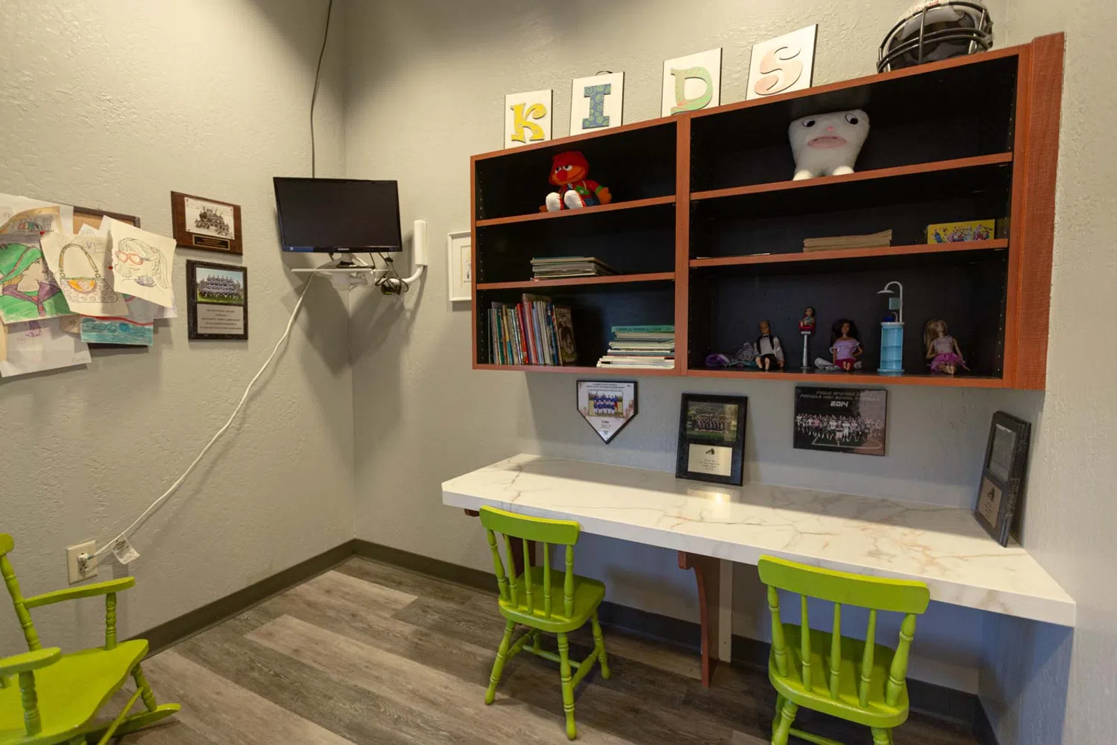 Children's study area with green chairs, wall-mounted TV, shelves with toys, books, and kids' artwork on the wall.