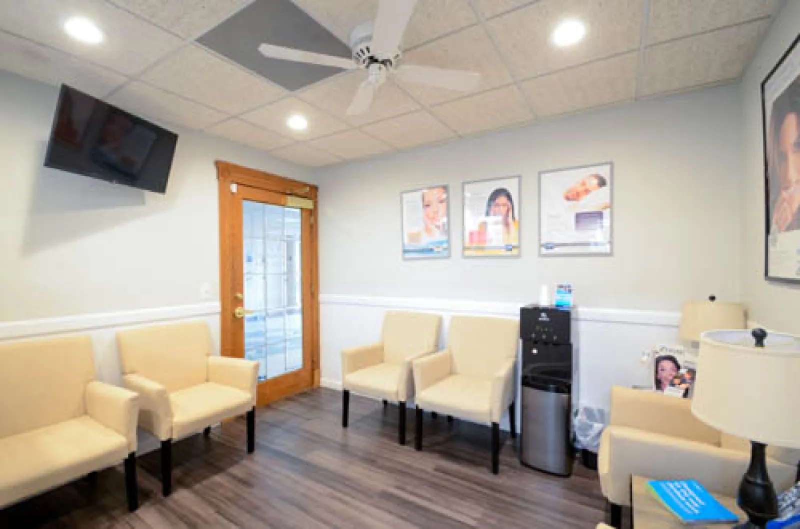 Bright waiting room with beige chairs, wooden door, water cooler, ceiling fan, and framed posters on light walls.
