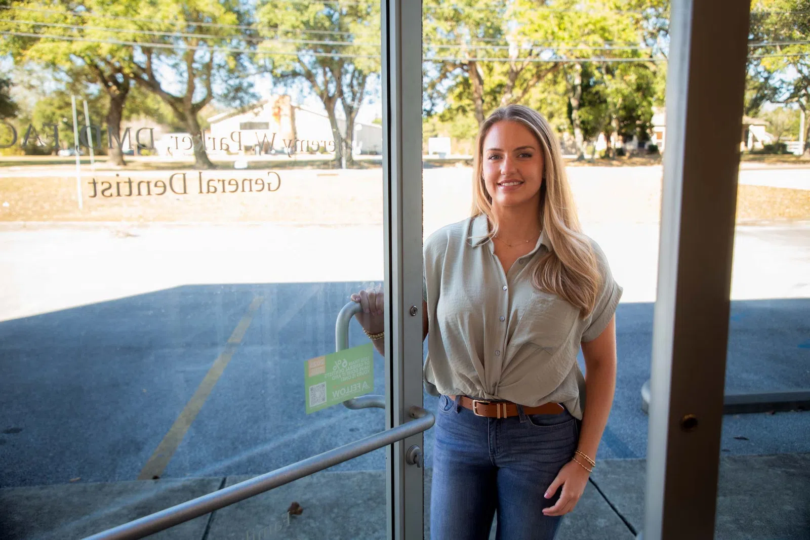 Smiling woman holding door open standing outside a dental office with trees and parking lot in background