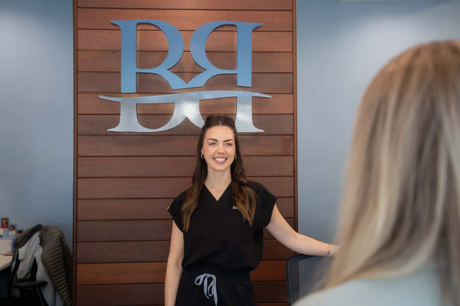 Smiling woman in black uniform standing in front of wooden wall with silver RRH logo in office setting