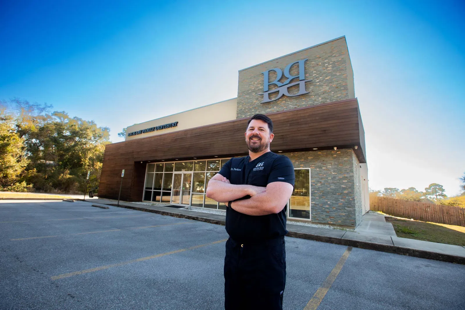 Smiling man in black scrubs stands with arms crossed outside modern dental office building under clear blue sky