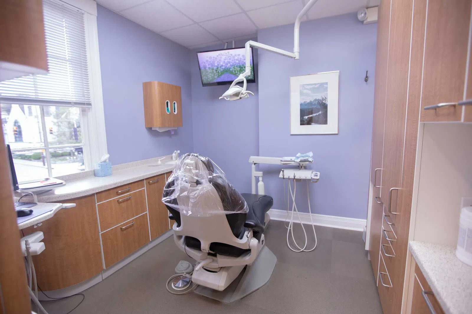 Modern dental office with patient chair, overhead light, cabinets, and monitor on soft purple walls under ceiling lights.