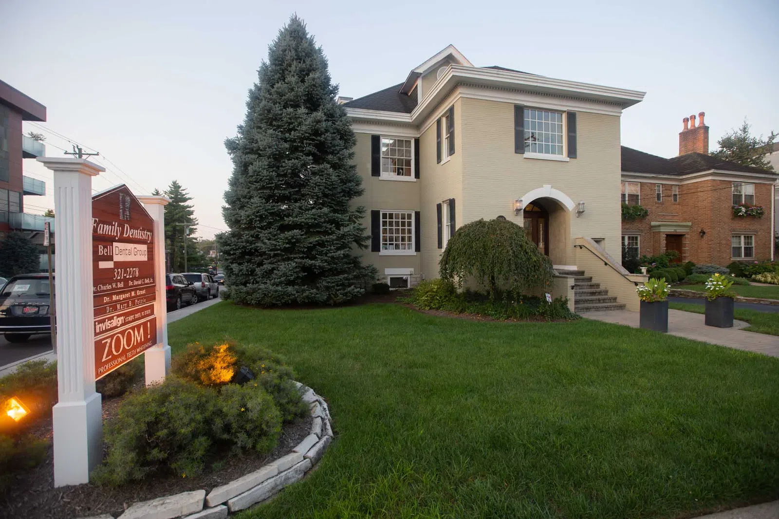 Two-story beige house with black shutters and a dental office sign in a landscaped green lawn.