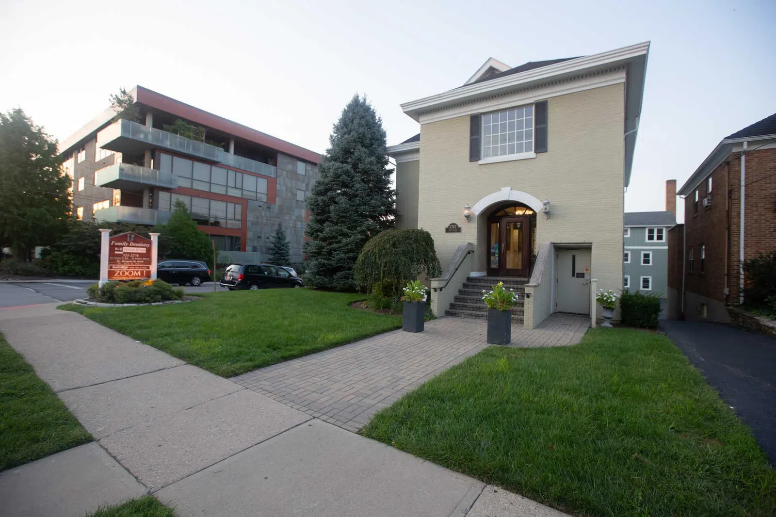 Two-story beige building with stairs, green lawn, sidewalk, trees, and modern apartment in the background.
