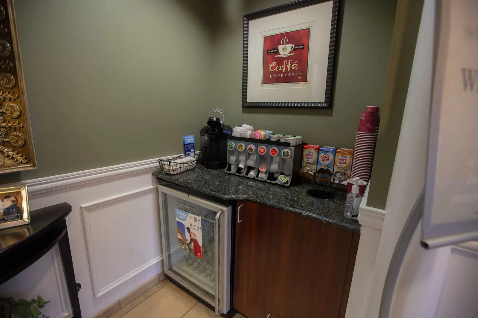 Small coffee station with espresso machine, condiment dispensers, cups, and mini fridge in corner of room.