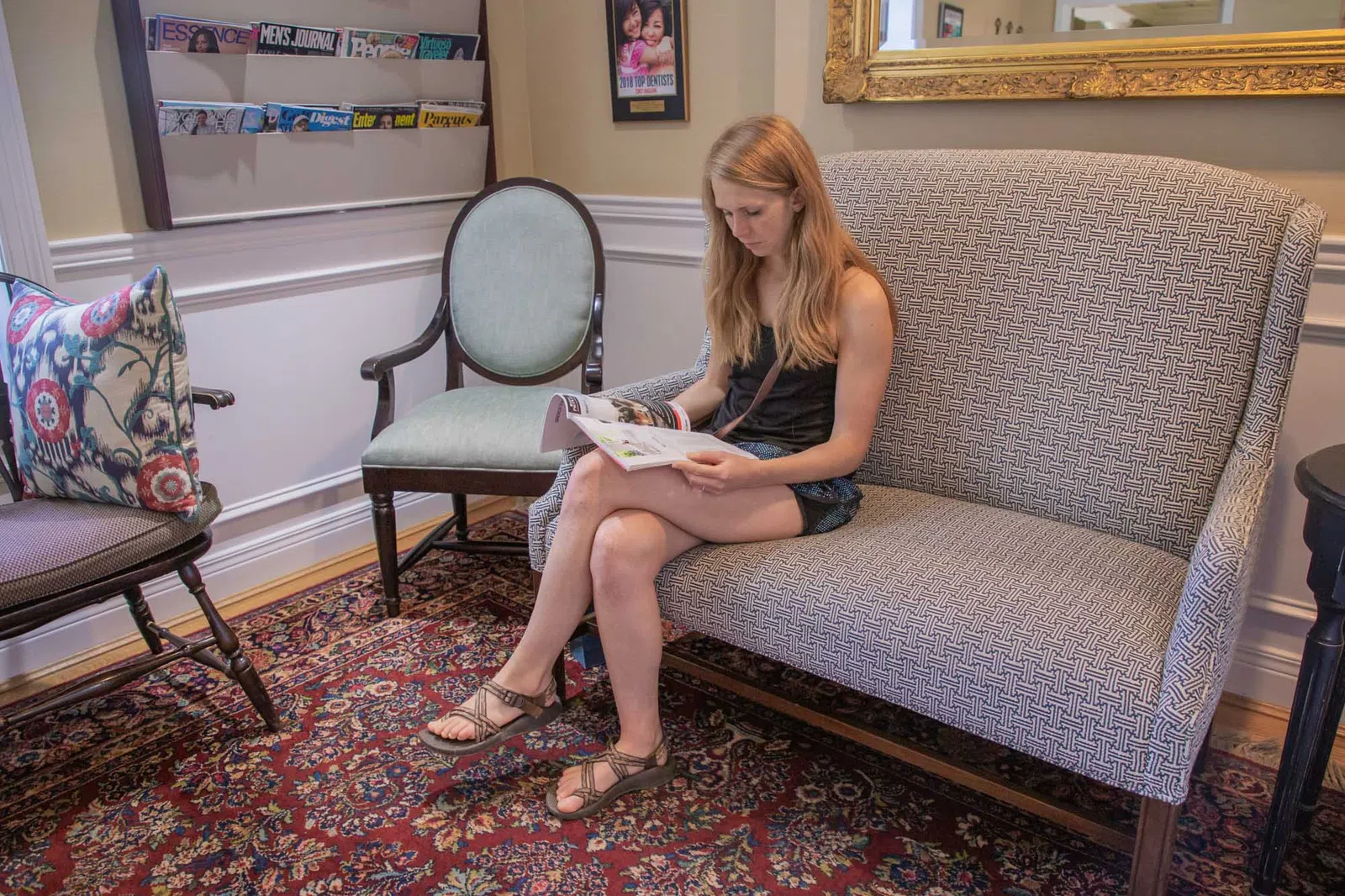 Young woman sitting on patterned sofa reading a magazine in a cozy waiting room with vintage furniture and colorful rug.