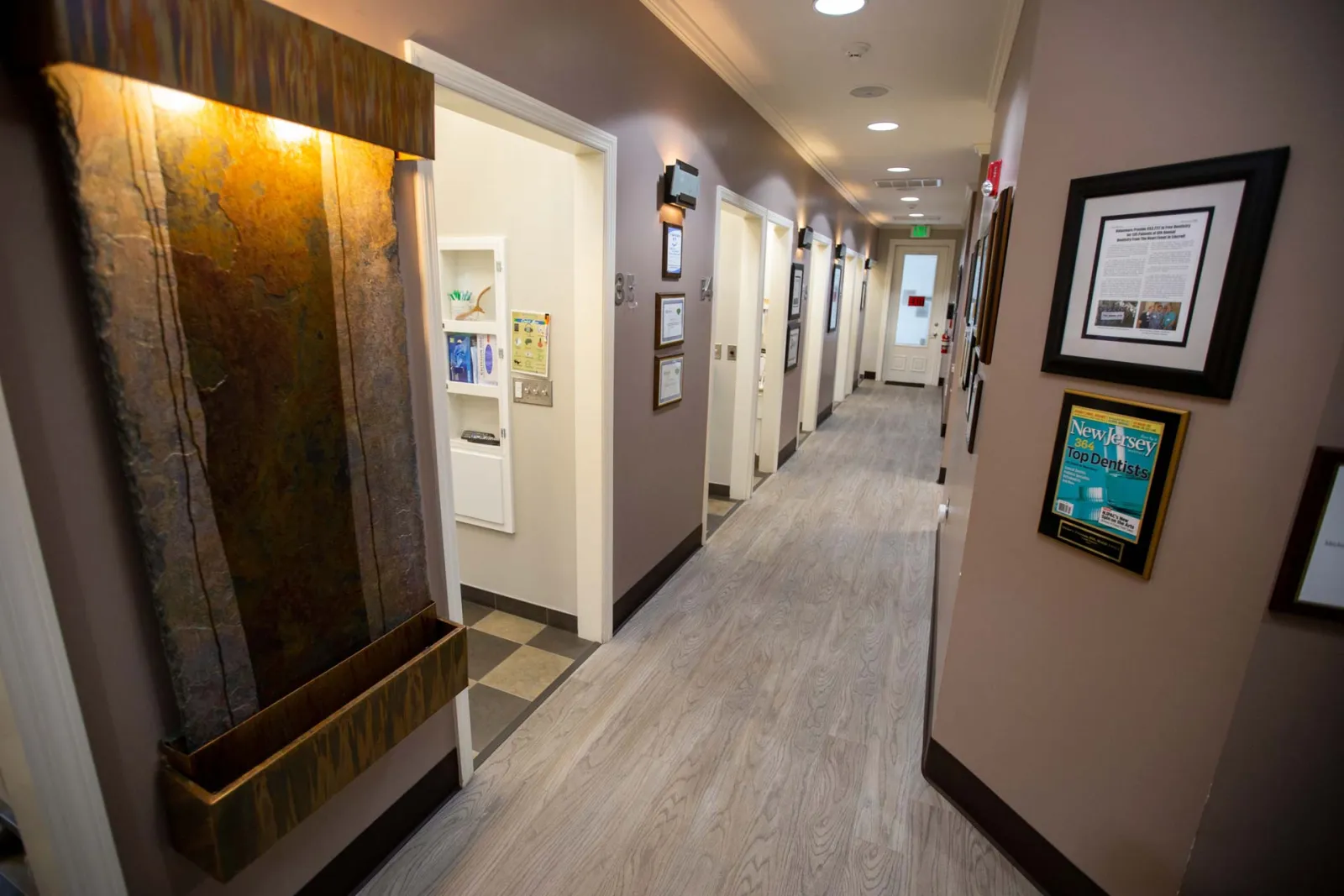 Well-lit dental office hallway with open patient rooms and framed awards on beige walls.