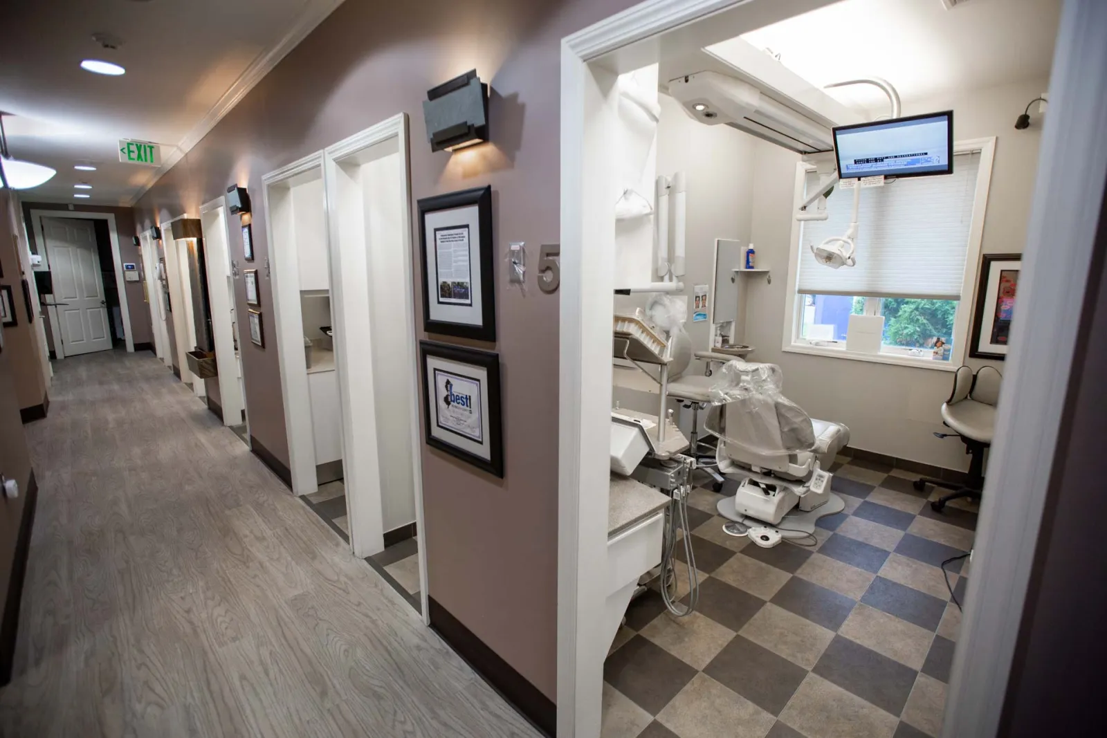 Bright dental clinic hallway with open patient rooms and modern dental equipment in one room visible on checkered floor.