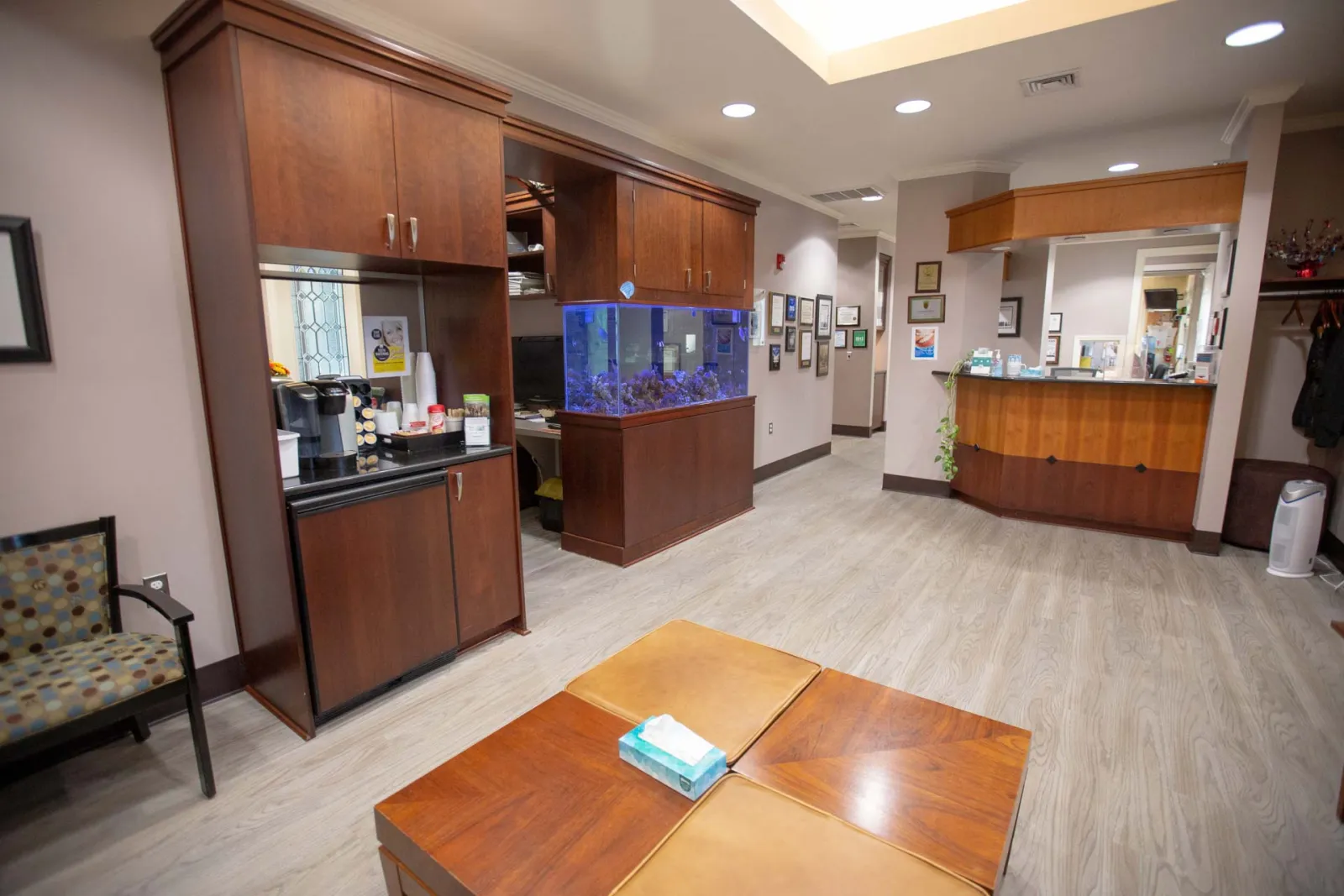 Waiting room with wooden furniture, fish tank, coffee station, and reception desk in a professional office.